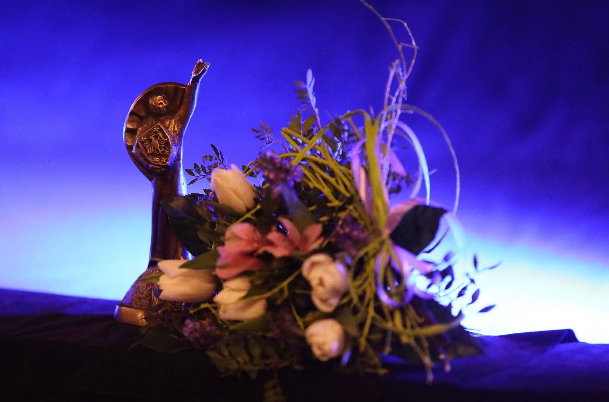 Na niebieskim tle statuetka metalowego Clemensa i bukiet kwiatów