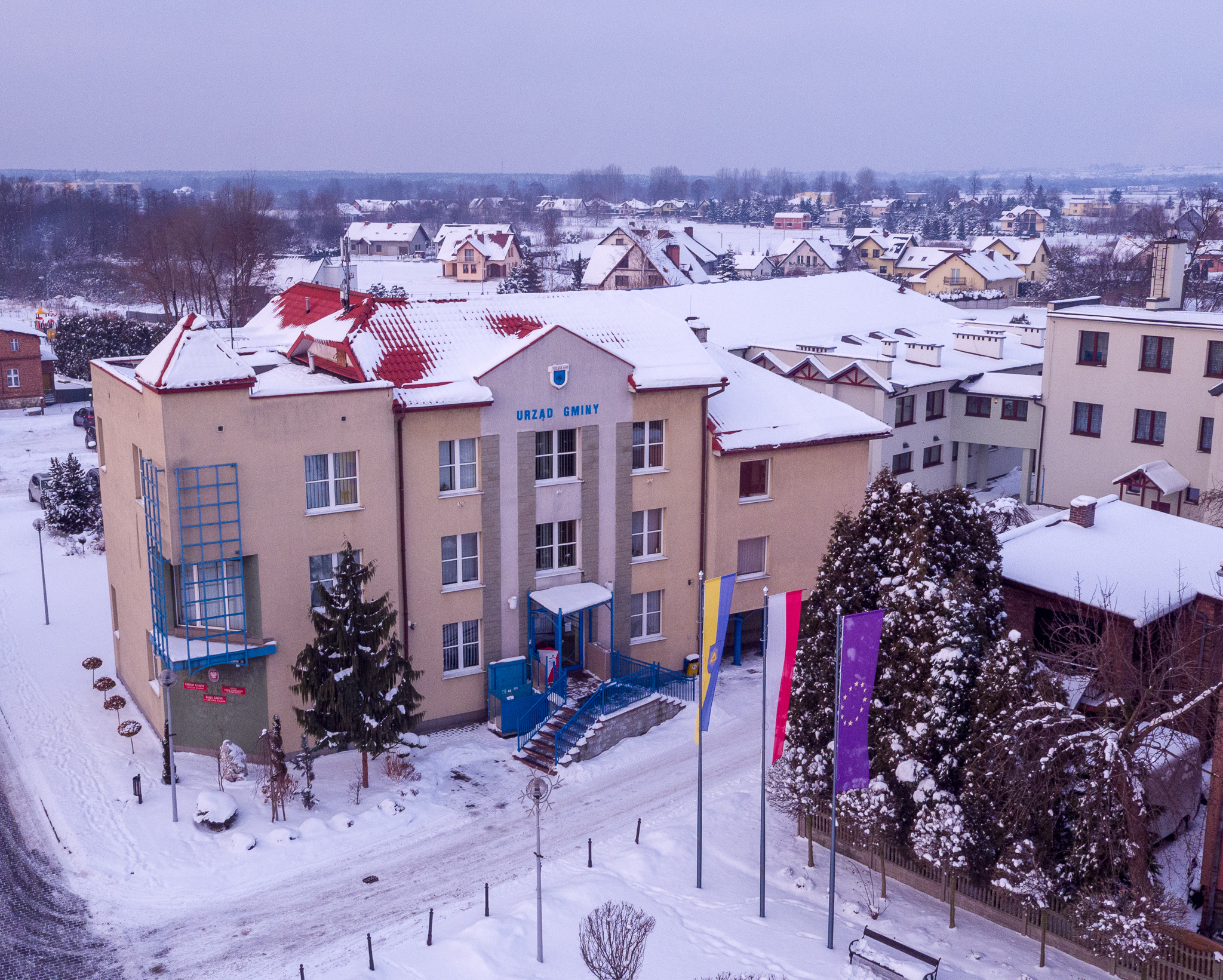 Zdjecie z drona. Na zdjęciu Urząd Gminy, flagi i okoliczne budynki mieszkalne.