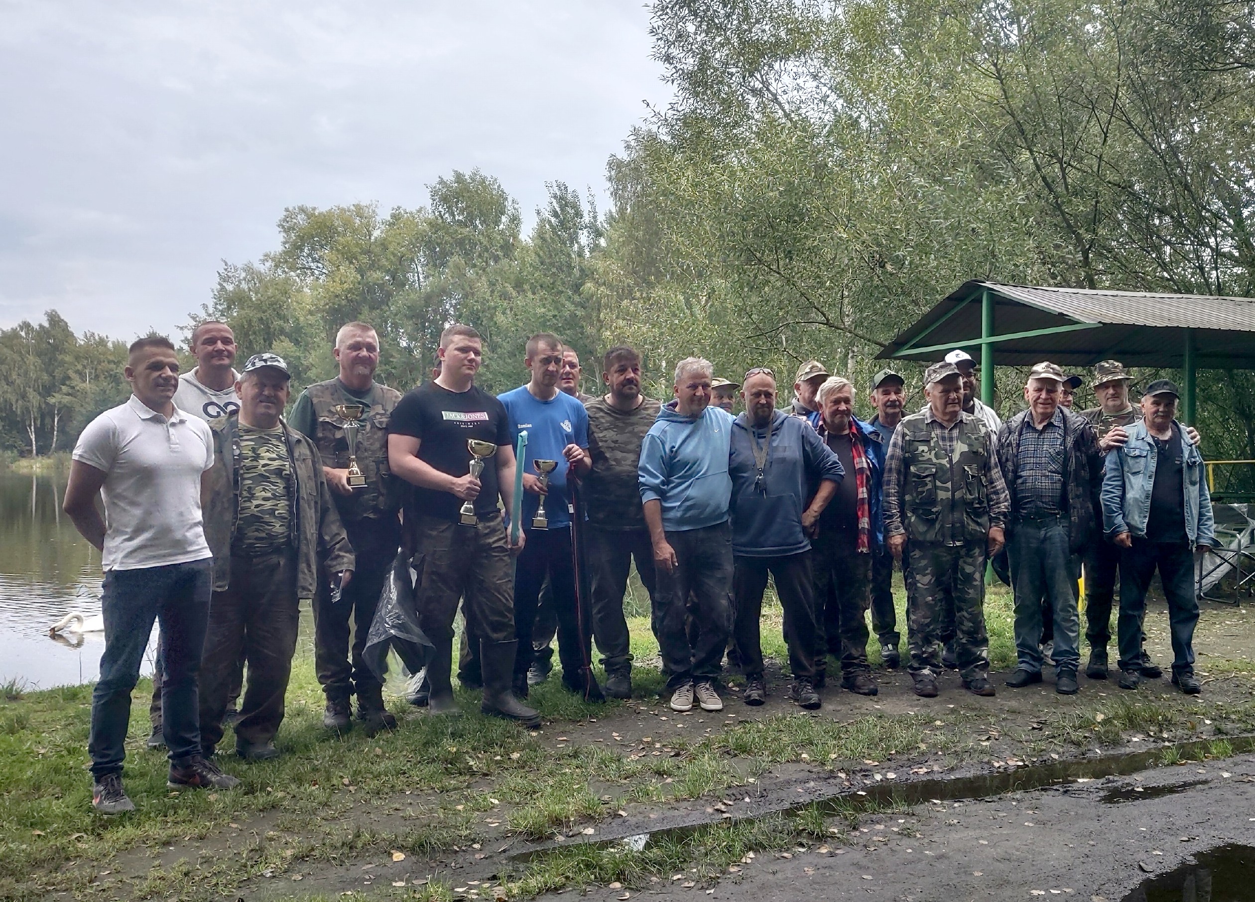 Mężczyźni pozujący do zdjęcia z pucharami na brzegach stawu. Po zawodach wędkarskich