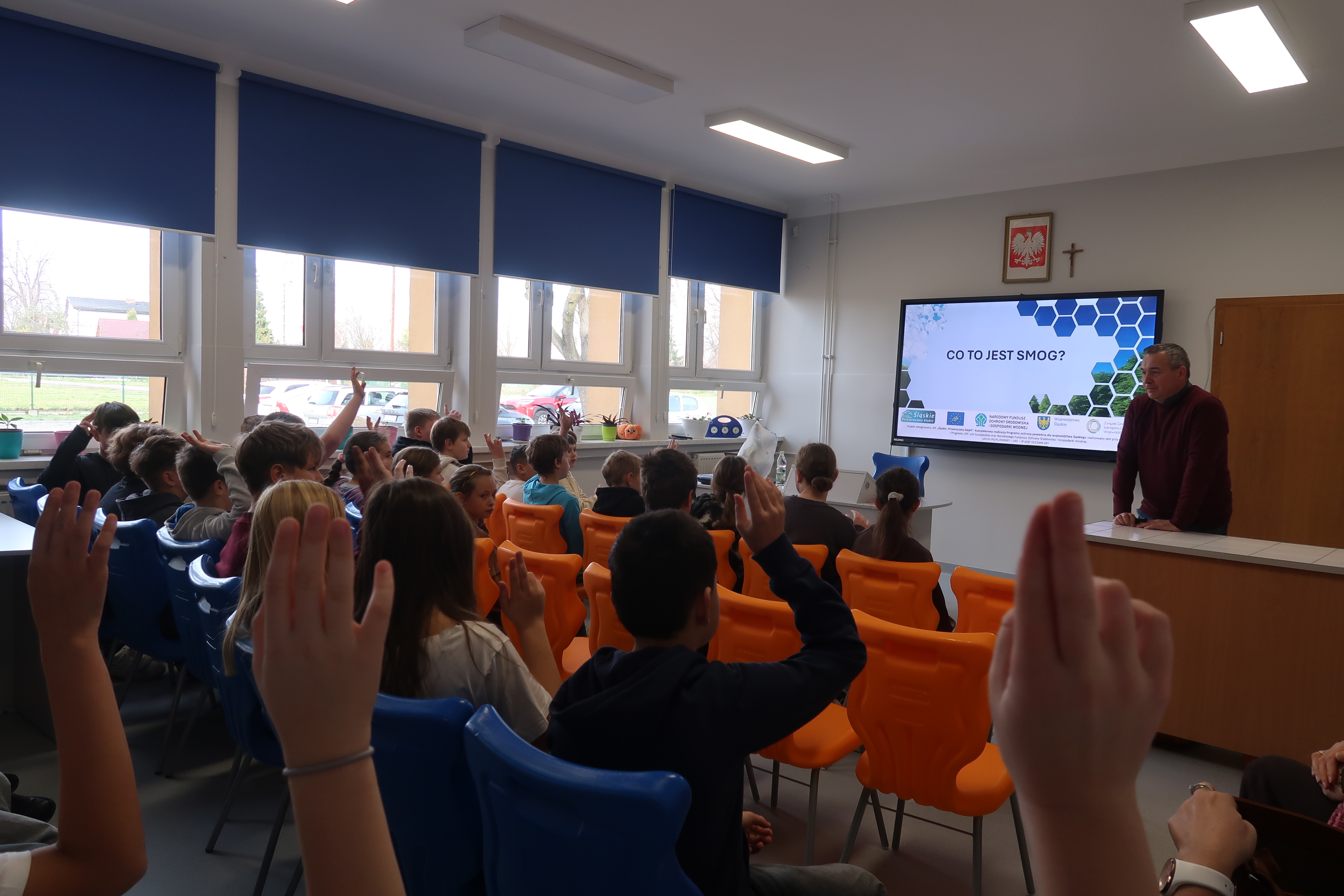 Dzieci siedzące w klasie przed multimedialnym ekranem. Zajęcia prowadzi mężczyzna. 